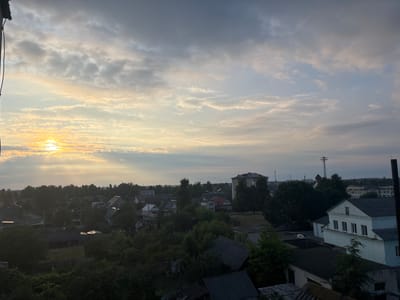 This is an aerial shot of a rural village in Belarus, likely during a hazy sunrise or sunset. The sun, partially obscured by clouds, casts a warm, golden light on the left side of the frame, illuminating the sky and creating a gentle glow. Wispy clouds stretch across the sky, some tinged with yellow and orange from the sun's rays.
The village below is a dense collection of houses, many with dark, sloped roofs, surrounded by lush green trees. Some of the houses appear to be made of wood, while others are brick. The overall impression is one of a peaceful, lived-in community. In the distance, larger buildings and a tall tower suggest a more developed area beyond the immediate village. The weather appears to be humid and possibly foggy, as indicated by the haziness of the light and the softness of the distant landscape. There are no visible people in the frame, and the activity suggested is simply the quiet life of a village. The lack of sharp detail due to the atmospheric conditions gives the image a serene and somewhat dreamlike quality.