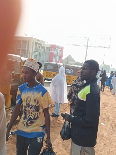 The scene depicts an outdoor urban or semi-urban environment in Gobirawa, Nigeria, observed during daylight. The immediate foreground is a reddish-brown dirt ground. The top-left corner is partially obscured by a light-colored object, possibly a finger or arm.

A young man with dark skin occupies the left mid-foreground, wearing a patterned beige and brown kufi cap, a blue and yellow graphic t-shirt, and dark trousers. He faces slightly left. To his right, another man with dark skin and a short beard wears a black jacket with a lime green and white shoulder stripe over light trousers. He holds a small dark object in his left hand and a black plastic bag in his right, looking right. A person in a brown garment is partially visible behind the young man.

In the midground, a person in a white flowing garment and light blue jeans stands with their back to the camera, positioned between two yellow motorized tricycles (auto-rickshaws). At least three such tricycles are visible on a paved or unpaved road. One tricycle has "ASAN" printed on its side. Further back on the right, additional indistinct figures are present, including one in a dark hoodie.

The background features multiple buildings under a bright, clear sky. A multi-story, light-colored structure labeled "SDS PLAZA" is on the left. To its right, a red and white building displays "ALHAM... CHICKEN." An empty metal billboard stands further right, and a conical roof structure is visible in the distance behind the buildings.