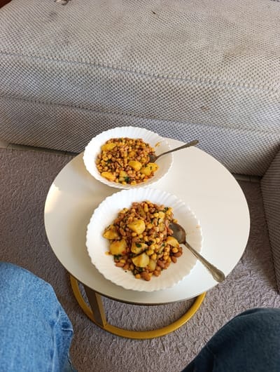 An indoor scene in Kahawa Wendani ward, Kenya, depicts a first-person perspective looking down at a meal. Two white fluted bowls, each containing a serving of Githeri (maize, beans, and potato pieces with green herbs), are positioned on a white circular side table with a gold-colored metallic base. A silver-colored spoon with a decorative handle rests in each bowl. The lower portions of a person's legs, clad in blue denim jeans, are visible in the foreground, indicating their proximity to the table. In the background, a large, light grey, textured sofa or upholstered furniture piece is visible, showing minor dark discolorations. The floor is covered with a light grey, textured carpet. The scene is brightly illuminated.