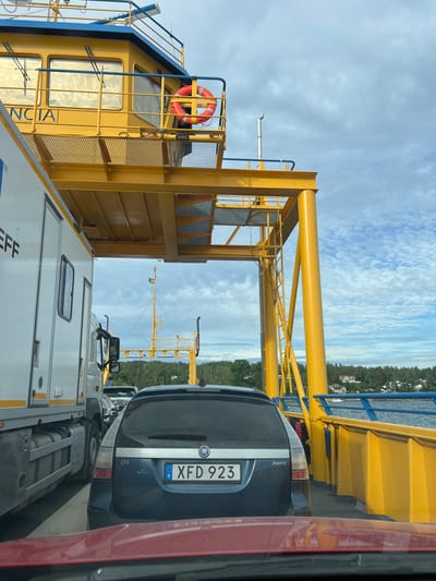 The image depicts a ferry filled with vehicles, with the perspective taken from inside one of the vehicles. The ferry itself is characterized by its bright yellow structure, including large support beams and railings, set against a backdrop of a partly cloudy sky and a body of water. On the left side of the frame, the superstructure of the ferry is visible, painted yellow with blue accents and the partially obscured word "NCIA" visible as text. A bright orange life preserver is attached to the railing.

Parked in a row are several vehicles. Closest to the viewer is the rear of a dark blue car, with its license plate clearly visible as "XFD 923" and a Swedish flag emblem. Next to it is a white truck, followed by other vehicles. The vehicles are lined up on what appears to be the deck of the ferry, suggesting they are either waiting to disembark or are currently in transit.

In the background, a tree-covered coastline with houses is visible, indicating a landmass near the ferry's route. The overall impression is one of travel and transportation across water, likely during daytime given the lighting. The weather appears to be fair, with scattered clouds and sunlight. The scene captures a common travel experience, possibly in a scenic coastal area of Sweden given the location context. No people are clearly visible within the frame.