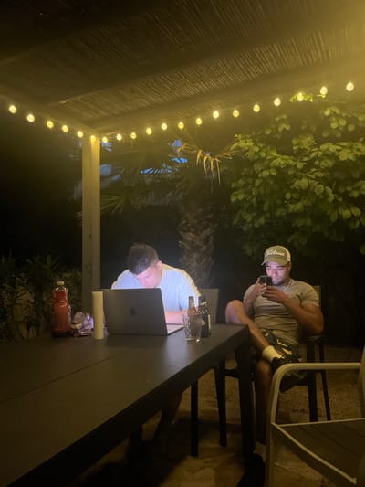 The image captures an outdoor scene at night, likely in a backyard or patio setting in el Poble Nou de Benitatxell / Benitachell, Spain. Two individuals are present: one is seated in a chair, looking at their phone, wearing a light-colored cap and a grey t-shirt. The second person is seated at a long, dark table, working on a laptop. Various objects are on the table, including a glass bottle and other containers. String lights are visible overhead, illuminating the area, and lush greenery surrounds the patio.