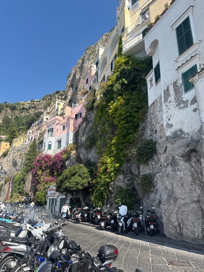 The image depicts a steep cliffside street in Pogerola, Italy, with colorful buildings clinging to the rock face. A clear blue sky dominates the upper portion of the frame, suggesting a sunny day. The buildings are painted in various pastel shades, including pink, yellow, and white, with green shutters on the windows. Lush green vegetation and vibrant purple bougainvillea cascade down the cliffs, adding to the picturesque scene.

Parked along the narrow, paved street are numerous scooters and motorcycles, indicating a mode of transportation common in this region. A few people are visible, including a person wearing a white jacket standing near a building entrance, and others on scooters. The scene appears to be set during daylight, likely late morning or early afternoon, given the strong sunlight and shadows. The overall atmosphere is one of a charming, bustling Mediterranean coastal town. There is some visible text on license plates of the scooters.