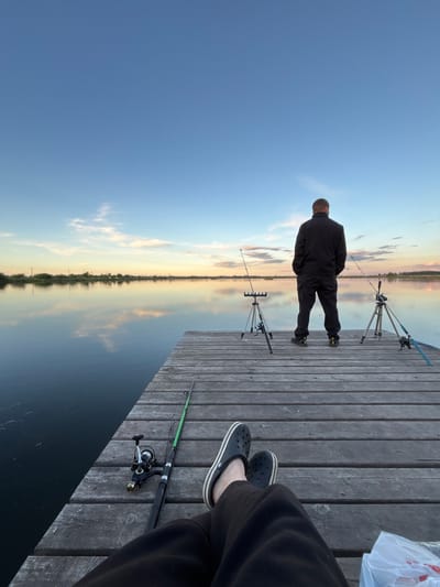 A pier scene with fishing activity is observed. Two individuals are present; one stands at the pier's edge facing the water, while another is seated nearby. Two fishing rod stands with fishing rods are visible near the standing individual. A fishing rod is also present near the seated individual. The pier appears to be made of wooden planks. In the background, the sky meets the water, creating a horizon with a sunset or sunrise effect. The location is identified as Khryashchevka, Russia.