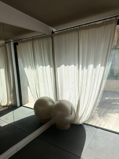 A well-lit gym space with a large mirror reflecting two white exercise balls. The room features floor-to-ceiling curtains, some of which are drawn back to reveal a wooden deck and greenery outside. The floor is a dark, textured mat. The overall impression is one of tranquility and focus on fitness. The image is captured in Montuïri, Spain.