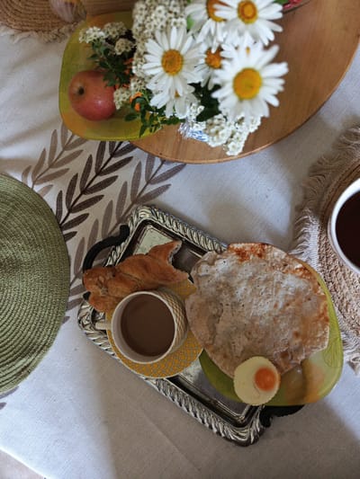 A top-down, close-up shot shows a breakfast setting on a table in Alexandrovskaya, Russia. A white tablecloth with a brown leaf pattern covers the table. A woven green placemat is on the left. In the upper portion of the frame, a wooden platter holds a small red apple and a bouquet of daisies and white wildflowers. In the lower portion of the frame, a silver tray displays a cup of coffee, a braided pastry, a piece of flatbread, and a small, circular piece of cheese with an orange yolk-like center. A woven coaster with fringe borders a white mug of dark liquid on the right.