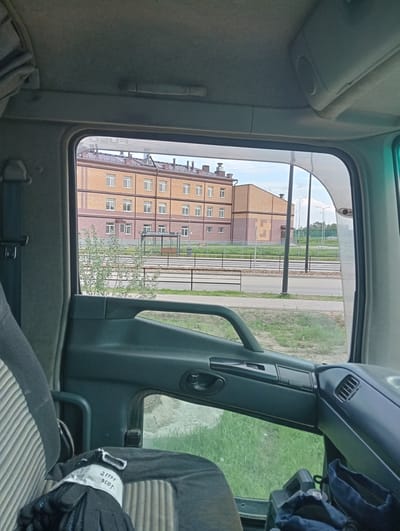 The image is taken from inside the cab of a truck, looking out the passenger side window. The window frames a view of a multi-story brick building with a pitched roof. The building appears to be an apartment complex or a school, with numerous windows visible. In front of the building, there is a fence, some trees, and a bus stop shelter. The foreground shows the interior of the truck cab, including a seat, a door handle, and some items on the seat. The overall scene depicts an urban or suburban environment with a focus on a building and its surroundings.