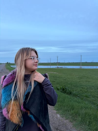 The image shows a woman with long blonde hair and glasses, standing outside in Azyakul', Russia. She is wearing a dark jacket with a colorful fur collar. In the background, there is a field of green grass, a flooded area, and power lines. The sky is overcast. The scene seems to be along a road or path on the edge of a field.