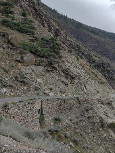 This image depicts a mountain road with a stone retaining wall and a culvert in Gushal, India. The road curves along the side of a steep, rocky mountain slope covered in sparse vegetation and scattered trees. The retaining wall, constructed from irregular stones, runs along the edge of the road, and a circular culvert is visible within its structure, suggesting it's designed to channel water drainage. The sky above is overcast. The scene is devoid of any people or animals.