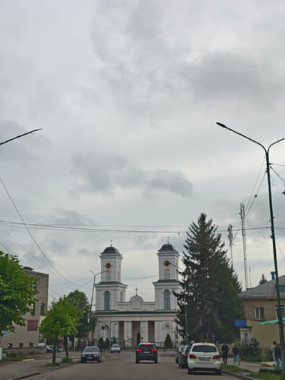 The image shows a street view in Nemyriv, Ukraine, facing the Holy Trinity Church. The church is light-colored with two bell towers, each topped with a cross and a clock face. A tall evergreen tree stands to the right of the church, partially obscuring the right tower. Several people are walking along the sidewalk on the right side of the street. Cars are parked or driving on the road. Overhead, streetlights with extended arms and power lines stretch across the scene. Cell phone towers are visible in the background. The sky is overcast. Buildings flank the street, including a multistory building on the left and a lower residential building on the right.