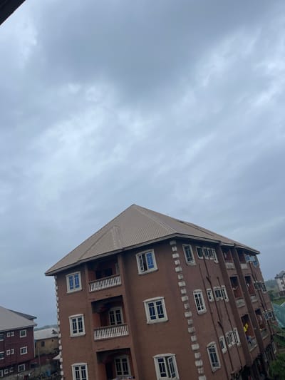 This is a medium-angle, eye-level shot of a multi-story apartment building in Iyiowa Odekpa, Nigeria. The building is constructed with a brown, stucco-like exterior and features a metal, sloped roof. Multiple windows with white frames and blue panes are visible on each level, some open and some closed. Balconies with white railings are also present. The sky above is overcast with gray clouds. To the left, another apartment building with a similar architectural style can be seen. The environment suggests an urban or semi-urban setting. There are no people clearly visible in the frame.
