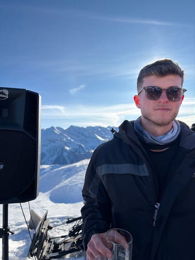 The scene is set in Schwendau, Austria, within a snow-covered alpine environment under a clear blue sky. A young adult male occupies the central-right portion of the frame, facing forward. He wears dark sunglasses with light-colored frames, a dark winter jacket featuring a grey chest stripe, and a patterned neck gaiter. His right hand holds a clear glass containing liquid residue, with partial text visible near the bottom. The sun, positioned behind his head, casts a bright backlight. To the left, a black RCF-branded professional audio speaker, also bearing a 'RIDING' logo, is mounted on a stand. Behind the subject and speaker, a panorama of snow-laden mountain peaks and valleys stretches into the distance. Below the speaker, partially buried in snow, is dark, folded mechanical equipment. No interactions with other individuals are depicted.