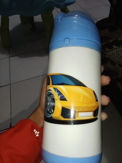 A white cylindrical bottle with light blue top and base sections is held by a human hand. The bottle features a printed graphic of a stylized yellow sports car, oriented towards the left, on its white central body. The hand, appearing from the right side, displays a dark skin tone and visible fingernails. A red long-sleeved garment with white text or embroidery, possibly "4jex", is partially visible in the lower left, indicating the person's left arm. The background consists of light-colored square floor tiles with dark grout lines. A light blue object with yellow, curved legs is partially visible in the upper left background, casting a shadow. A dark, patterned fabric object is present in the far right background. The specific location within Inxuba Yethemba Local Municipality, South Africa, cannot be determined from the visual information provided.