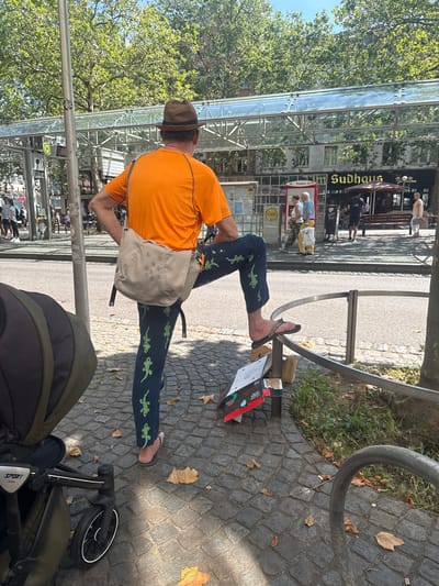 The image depicts a man standing on a cobbled sidewalk in Bonn, Germany. He is wearing a brown fedora, an orange t-shirt, and dark blue pants adorned with a repeating pattern of green lizards. A beige messenger bag is slung across his body. His right leg is bent and resting on a metal railing, while his left foot is in a black flip-flop. In the foreground, a dark green stroller is partially visible to the left. Behind the man, a glass-enclosed structure with metal framing stands, likely part of a public transportation station or market. A sign with the word "SUDHAUS" is visible on a building behind the glass structure. The environment is urban, with trees lining the background and a street visible to the right. The ground is paved with interlocking stones, and scattered autumn leaves are present. The overall impression is a casual outdoor scene on a sunny day.