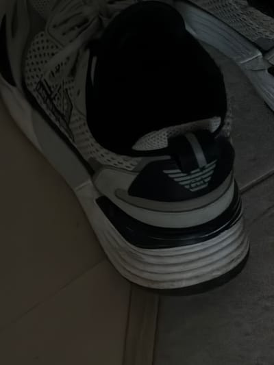 The image shows a close-up, low-angle view of a black and white sneaker. The sneaker is partially obscured by shadow, but the sole appears to be made of layered, ribbed material in white and black. The side of the sneaker features a dark navy blue section with the Emporio Armani eagle logo in a lighter shade. A grey stripe is visible above the logo. The upper portion of the sneaker is made of a white mesh material with black accents. The background is dark and appears to be a tiled surface, possibly a floor. The image does not contain any discernible elements that would suggest a location in Crokhovo, Russia, or any specific activity.