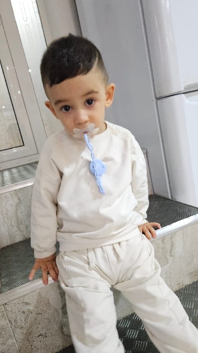 An image taken indoors in Vazovo, Bulgaria, depicts a light-skinned male child, approximately 2-4 years old, positioned centrally. He has short, dark hair with faded sides and a prominent part on the left. The child wears a cream-colored long-sleeved shirt and matching pants. A clear, flower-shaped pacifier with a white nipple is in his mouth, connected to a light blue, braided pacifier clip secured to the front of his shirt. The child is in a semi-sitting position on a step or ledge, which features a light-colored surface with a darker, textured metallic or ridged tread. His hands are resting on the step surface to his sides. In the background, a white-framed window with vertical elements is visible on the left, and a section of a white, two-door appliance (likely a refrigerator) is visible on the right. The child's eyes are directed forward.