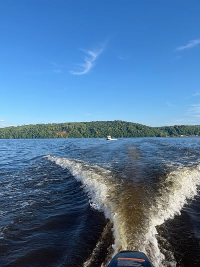 The image depicts a scenic view from a boat moving through a body of water, likely a river or lake, with a densely forested shoreline. The perspective is from the stern of the boat, looking towards the left and slightly forward. The water is dark blue and disturbed by the boat's wake, creating foamy white crests and ripples.
The shoreline is covered in a thick, green forest that extends to the horizon. A clear, bright blue sky with a few wispy white clouds dominates the upper left portion of the image. The sun appears to be high in the sky, suggesting it is daytime, likely mid-morning or afternoon, with good weather.
A small white boat is visible in the distance near the shoreline. The motor of the boat from which the photo is taken is partially visible in the lower right corner, showing a blue housing with some orange accents and text that is mostly illegible due to glare and focus.
The overall mood is peaceful and serene, capturing a moment of leisurely travel through a natural landscape. The location is identified as Siukterka, Russia.