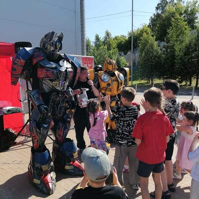 An outdoor event is depicted in Трубачеевка, Russia, under clear blue skies, featuring costumed performers and several children. In the foreground, a detailed, life-sized costume resembling Optimus Prime from the Transformers franchise stands prominently on a paved surface composed of light-colored pavers. This costume is predominantly dark gray and black, accented with red and blue armor panels, featuring stylized red flame patterns and illuminated blue lights on the chest and lower legs. A white electrical cord extends across the ground near its feet. To the right and slightly behind Optimus Prime, a person in dark attire, wearing glasses, holds a microphone to their mouth with their right hand and a silver, handheld device in their left. Adjacent to this individual, a second detailed costume, resembling Bumblebee, stands. This costume is primarily yellow with black accents and features visible headlights. Behind the performer and Bumblebee, a white banner or sign displays the red Autobot symbol.
A group of approximately eight children, ranging in age, are gathered around the performers. One girl with braided hair, wearing a pink shirt and patterned shorts, is extending her hands towards Bumblebee. Next to her, a boy in a black and white patterned shirt also has his hands raised towards Bumblebee. A boy in a red t-shirt and dark shorts stands with his back partially to the camera. Another boy, with his back completely to the camera, kneels or crouches on the ground, wearing a gray baseball cap and holding a yellow object. Further to the right, a girl with face paint (red and white design on her cheek) wears a light pink dress.
To the far left, a red canopy or tent structure is visible, with a small black table underneath holding a white folded cloth. The ground is paved with rectangular pavers, on which faint white chalk marks are visible. In the background, a large, light-colored building with distinct horizontal lines or panels rises on the left side. The background to the right consists of numerous green trees and dense foliage, with a utility pole and overhead power lines also present. A paved road or path is visible behind the children on the right side of the frame.