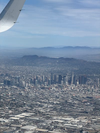 An aerial, high-angle, vertical shot captures a sprawling urban landscape, likely Los Angeles, as seen from an airplane. The foreground is dominated by the left wing of the aircraft, its white surface detailed with rows of small circular indentations and two small black fins protruding from its edge. The wing curves gracefully into the upper left quadrant of the frame, against a hazy blue sky filled with thin, wispy clouds.

The mid-ground reveals a dense cityscape, a vast expanse of buildings stretching towards the horizon. Numerous skyscrapers, characterized by their rectangular forms and varying heights, form a prominent cluster, suggesting a downtown area. Interspersed with these tall structures are a multitude of smaller buildings, creating a textured urban fabric. Roads and highways weave through the cityscape, indicating a well-developed transportation network.

Further in the distance, the city gives way to rolling hills and then to a range of larger mountains, their forms softened by atmospheric haze. The landscape suggests a city situated in a basin, surrounded by natural topography.

The lower portion of the image shows a densely packed industrial or commercial zone, with many large, flat-roofed buildings, likely warehouses or distribution centers, covering a significant area. These structures are arranged in a grid-like pattern, with roads and parking lots visible between them. This area could be indicative of a part of Huntington Park, known for its industrial and commercial activity, given its proximity to the downtown Los Angeles core visible in the distance.

The overall scene depicts a vast, complex urban environment under a clear, albeit slightly hazy, sky. No people are discernible in the image, and the focus is on the urban development and the natural surroundings.