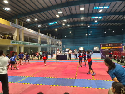 The image shows an indoor sports arena, likely a gymnasium, with a bright red and blue mat laid out for a game. Several young women are actively playing on the mat, dressed in blue and red sports uniforms. Some are in mid-action, while others appear to be observing. Spectators are visible in the background, some seated in bleachers, and others standing. There are banners and signage around the arena, indicating a sports meet. One prominent banner reads "KENDRIYA VIDYALAYA SANGATHAN SPORTS MEET 2026". The architecture of the building, with its high ceiling and metal beams, suggests a large public indoor facility. The lighting appears to be a mix of fluorescent and natural light from skylights. Based on the provided information, this scene is located in Phagwara, India, within the Kendriya Vidyalaya Sangathan Sports Meet.