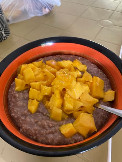 A close-up overhead shot shows a bowl of arroz con leche, a rice pudding, topped with diced mangoes. The bowl has an orange interior and a black exterior rim. A spoon is partially visible on the right side of the bowl. The bowl sits on a beige, tiled table. In the background, to the left, is a black and white patterned object and a clear plastic bag containing what appear to be loaves of bread. The scene is set indoors, likely in a residential kitchen or dining area, within the city of Bacong, Philippines.