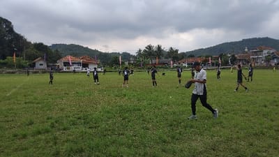 The image depicts an outdoor sports field scene in Panjalu, Indonesia, under an overcast sky. A group of approximately fifteen individuals, mostly young men or teenagers, are spread across a wide, green grassy field. Most of them are wearing dark-colored (black or dark grey) sports jerseys, some featuring white numbers such as '18', '6', and '8'.
In the foreground, a person wearing a grey and white striped shirt, dark pants, and a dark cap walks towards the right side of the frame, holding a dark glove or small bag in one hand. The other individuals on the field appear to be standing or moving casually, suggesting a break in activity, a warm-up session, or an informal game. The field shows some faint markings, and a partial goalpost structure can be seen in the distance on the left side, indicating a potential football (soccer) pitch.
The background reveals a rural or semi-rural setting. Several houses with distinctive tiled roofs and open structures are visible, nestled among abundant green trees and dense foliage. Rolling, tree-covered hills stretch across the horizon, framing the scene beneath a cloudy, grey sky. The lighting is diffused, consistent with an overcast day or possibly late afternoon. The overall impression is one of a relaxed, informal gathering on a local sports ground.