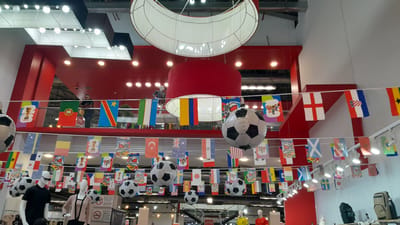 An indoor scene displays a vibrant, festive atmosphere, likely a retail space or event venue, decorated with numerous national flags and oversized inflatable soccer balls. The primary color scheme features prominent red accents in the architecture, contrasting with white ceilings and grey walls. A large, crescent-shaped white structure hangs from the ceiling, and a red cylindrical object is also visible overhead.
The flags, strung in multiple rows, represent various countries, suggesting a theme related to international sports or events. Some flags are clearly identifiable, such as Portugal, DR Congo, Colombia, England, Croatia, Ghana, Australia, Turkey, and Brazil. Small pictures or logos are also integrated into some of the flag banners.
Several large, black and white inflatable soccer balls are suspended in the space, adding to the sports theme. In the foreground, mannequins are displayed, dressed in casual attire and sporting accessories. One mannequin wears a black t-shirt and sunglasses, while another is dressed in a light pink cardigan and a white top.
In the background, a person is visible standing on an upper level, looking out over the decorated area. The lighting appears to be a mix of overhead fluorescent lights and spotlights, creating a well-illuminated environment. The overall impression is one of a lively and engaging space, designed to evoke excitement and national pride, possibly for a sporting tournament. The location is identified as Unknown, Venezuela, though the specific type of establishment is not explicitly stated.