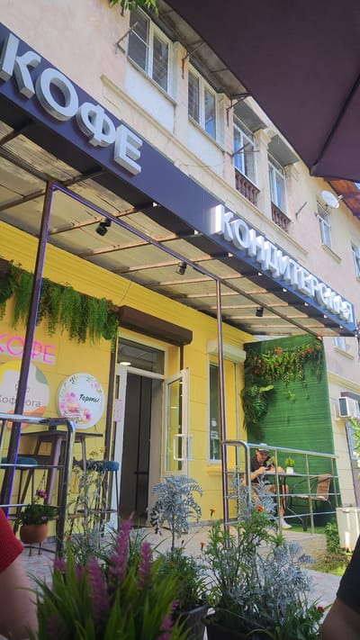 The image shows the exterior of a cafe with a bright yellow facade. A large sign above the entrance reads "КОФЕ" in white, three-dimensional letters on a dark blue background. To the right, another sign says "КОНДИТЕРСКАЯ" in similar white lettering.

There are several round signs on the yellow wall. One, illuminated in pink neon, says "КОФЕ". Another, with a floral design, has the word "Торты" (Cakes) written on it.

In front of the cafe, a covered outdoor seating area is visible. Potted plants with green and purple foliage are placed in the foreground. A person is sitting at a small table on a patio to the right, seemingly using their phone. The area appears to be well-lit, suggesting it is daytime, and the presence of plants and outdoor seating implies pleasant weather.

The setting appears to be a street-level establishment in a town or city, possibly in Dagomys, Russia, given the context. The overall impression is of a welcoming cafe with a focus on coffee and pastries.