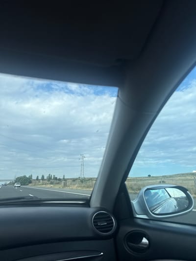 The image captures a view from inside a car traveling on a highway, with the sky and roadside scenery visible. The car's interior dashboard, a side mirror, and a portion of the windshield are prominent in the foreground. The side mirror reflects the road ahead and the sky. The exterior view shows a cloudy sky with scattered patches of blue. A power line pylon stands tall on the right side of the highway, with trees and a fence lining the road. Several cars are seen on the highway in the distance. The landscape appears to be dry and grassy with a few scattered trees. The image does not provide any information that would allow for identification of the location in the city of Zheleznik, Bulgaria.