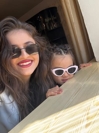 This image depicts an adult female and a young child in an indoor setting in Goris, Armenia. The adult female, with long, wavy dark brown hair, occupies the left side of the frame, facing slightly right and smiling broadly. She wears dark, hexagonal-framed sunglasses and red lipstick, revealing white teeth. A light-colored garment is visible on her left shoulder. To her right, a young child, approximately 3-5 years old, peeks over a light-colored wooden surface, resting both hands on it. The child's dark hair is styled with multiple small braids or ponytails secured with pink or light-colored bands. They wear white or light pink cat-eye shaped sunglasses with dark lenses and are looking slightly downward and to the right with a partially open mouth, exhibiting a playful demeanor. The wooden surface extends diagonally across the lower right portion of the frame, brightly illuminated by sunlight, highlighting its grain. In the background, to the left, a dark cabinet or shelving unit contains bottles or glassware. To the right, a light-colored curtain and a textured wall are visible, indicating a window area. The scene captures a shared, cheerful moment.