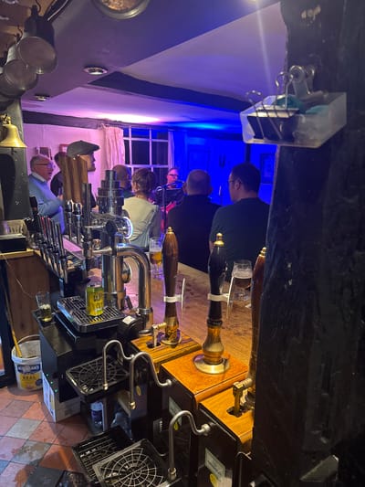 The image shows the inside of a pub in Babergh, United Kingdom, specifically behind the bar. Five people stand at the bar, appearing to be customers. A series of beer taps are visible along the bar top, with glass holders underneath. In the background, there is a window with multiple panes. The lighting is a mixture of blue and violet. A bucket labeled "HELLMANN'S REAL" is visible on the floor behind the bar. The floor is tiled with an alternating pattern of light and dark squares.