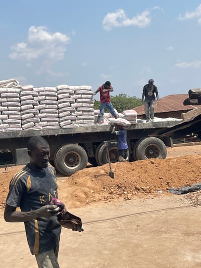 Several laborers load a truck with stacked bags of cement under a bright sky. One man in a red plaid shirt stands atop the bags, handling the load. Another man in blue clothes appears to be assisting from the side, with a shovel nearby. A third laborer is seen standing above the truck bed. The man closest to the camera is wearing a dark shirt and wiping his face. The scene suggests manual labor in construction or supply activities.