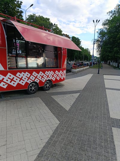 The image depicts a red food truck parked on a paved sidewalk in Gubkin, Russia. The food truck has a white geometric pattern on its side and an open red awning. A menu is visible through the truck's windows, displaying text in Russian. Street lamps and trees line the sidewalk, and several cars are parked on the street behind the food truck. The sky is partly cloudy. The scene appears to be a public space, possibly a park or a pedestrian area.