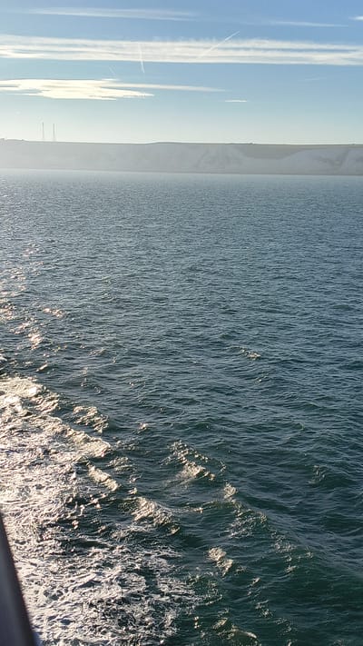The image depicts a view from a vessel sailing in the English Channel, with the White Cliffs of Dover in the background. The sea is a deep blue, with sunlight glinting off the ripples and waves. In the foreground, a wake of white foam trails behind the vessel. The White Cliffs, characteristic of Dover, are visible as a light-colored, sheer escarpment. Two tall radio masts are prominent on the clifftop. The sky above is a clear blue, with a few wispy white clouds and contrails from aircraft. The overall impression is one of a bright, sunny day at sea.