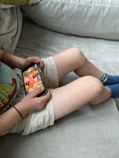 A young child, wearing beige shorts and a red, blue, and yellow t-shirt, sits on a gray couch, holding a black smartphone in both hands, watching a cartoon on the screen. The child is wearing a blue and yellow sock on the right foot. The child is sitting in a relaxed manner, with legs extended and feet on the couch. The image was taken indoors, likely in a living room. The location of the image is Rye, New York, United States. The coordinates are 40.952 latitude and -73.708 longitude.