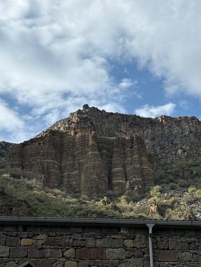 A geological formation is visible next to a building in Goght, Armenia. The formation is characterized by layered rock formations and vegetation. The building has a stone exterior with a metal pipe or gutter running along its side. The sky above has a mix of blue and white clouds.