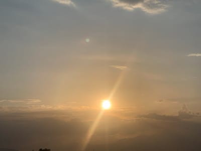 The image captures a picturesque sunset over a mountainous landscape, likely in Somoniyon, Tajikistan. The sky is a gradient of soft oranges and yellows, transitioning to a muted blue at the top, indicating the approach of evening. The sun, a bright, glowing orb, is positioned in the center-right of the frame, casting a strong, diagonal beam of light that cuts across the scene.
Wispy clouds are scattered throughout the sky, some catching the golden light of the setting sun, appearing as soft, luminous brushstrokes. Below the sky, the silhouettes of mountains are visible, their dark forms providing a dramatic contrast to the illuminated sky. In the foreground, the dark silhouette of trees suggests a natural or semi-rural setting.
The overall mood is serene and tranquil, evoking the peacefulness of dusk. The presence of the sun and the atmospheric conditions point to a clear or partly cloudy evening. There are no discernible people or man-made objects that are prominent in the scene. The image is devoid of any visible text.