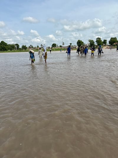 A group of people are wading through a wide, shallow river under a cloudy sky. Many of them are carrying items on their heads, including what appear to be large containers and sacks. The riverbanks are lined with trees and small buildings, suggesting a rural or semi-rural setting, likely in Kunini, Nigeria, given the context. The water is murky brown and appears to be flowing gently. The people are dressed in casual clothing, with some wearing traditional attire. The overall scene depicts a daily activity of crossing the river, likely for transportation or to access resources.