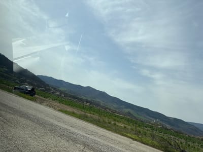 The image shows a landscape with a road running diagonally across the upper right. Below the road is a green field that transitions into mountains in the distance. Above the road is a sky with clouds. The reflection of the interior of a car is visible at the bottom of the frame. The location is likely near Dzhurovo, Bulgaria, given the user's prompt. A car is parked on the side of the road. The vegetation is consistent with a rural setting. A contrail from a plane is visible in the sky.