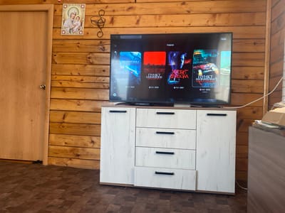 The image shows a television screen displaying movie posters in a room with wooden paneled walls. A door is visible on the left side of the image, and a white dresser with drawers is positioned beneath the television. Above the television, an icon of the Virgin Mary and child is mounted on the wall. The scene is indoors, and the overall aesthetic suggests a rustic or cabin-like interior, which aligns with the provided location information of the village of Kulchum, Russia.