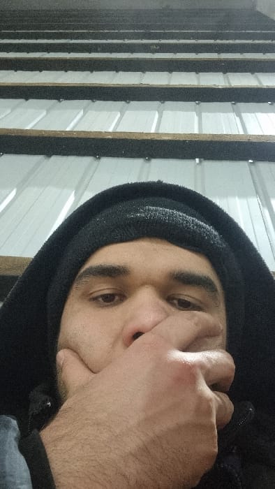 A close-up, rotated view shows a man in a black hoodie with a matching beanie. He is gripping his face with his hand. His eyes are open, and his expression is obscured by his hand. The background features vertical lines of white corrugated metal panels interspersed with dark wooden or composite strips. The lighting is somewhat dim, creating shadows and highlights across his face and the textured wall.