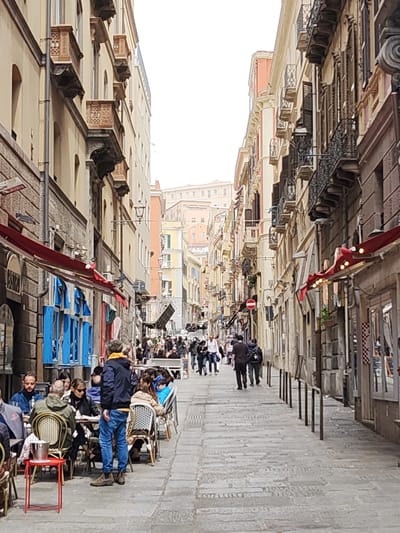 The image depicts a narrow street in Cagliari, Italy, populated with people and lined with buildings. The street is paved with stone blocks. On the left side, there is a restaurant with outdoor seating where people are seated at tables, eating and talking. The buildings on both sides of the street are multi-storied with balconies and various architectural details, including shutters and awnings. A group of people is walking further down the street, which narrows into the distance. A red "no entry" sign is visible on the right side of the street. The sky is overcast, casting a soft light on the scene.