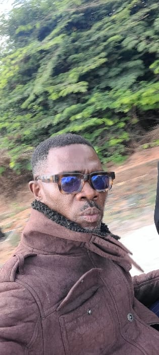 A dark-skinned male individual is depicted in a medium-close-up, appearing to be in motion within the city of Alayi, Nigeria. The specific location within Alayi is not discernible from the visual evidence.
The man, appearing middle-aged, has short, dark hair with a faded cut on the sides. He has a mustache and short facial hair around his mouth and chin. He is wearing large, tortoise-shell framed sunglasses with reflective blue lenses, which obscure his eyes. His mouth is slightly pursed.
He is dressed in a dark brown, possibly suede or textured fabric, jacket that features a prominent black faux fur collar. The jacket also has shoulder epaulets and flap pockets visible on the chest area. A dark, indistinct object, possibly part of a vehicle or another person, is partially visible on the far right.
The background consists of blurred green foliage, indicative of trees or bushes, directly behind the subject, with a lighter, indistinct strip below the foliage. The significant horizontal motion blur in the background suggests the photograph was captured while the subject or the camera was moving.