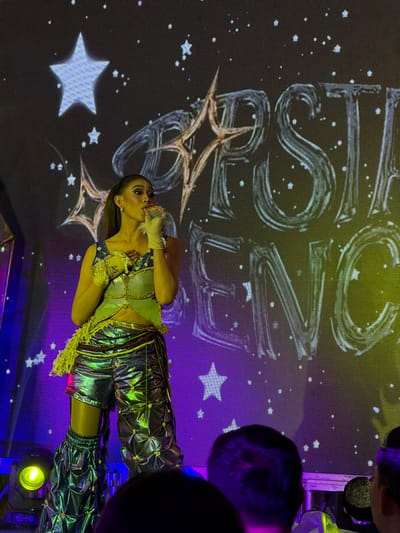 A woman stands on a stage, holding a microphone to her lips and her hand on her chest, appearing to be singing. She is wearing a futuristic, metallic outfit consisting of a sleeveless, embellished top and baggy, textured pants. The background features a large screen displaying a starry night sky with the words "APESTA ENON" written in a stylized font. The stage is lit with purple and yellow lights, and spotlights are visible on the left. The lower part of the image shows the backs of several audience members' heads, with one person on the far right. The scene suggests a live performance, possibly a concert or a show. The style of the text and the performance attire suggest a modern or theatrical event. While the image doesn't directly provide the location, "APESTA ENON" could be a reference to a festival or event, and a general search indicates that "Apexta Enon" was a title associated with a show by a Filipina singer who performed in Cebu City, Philippines.