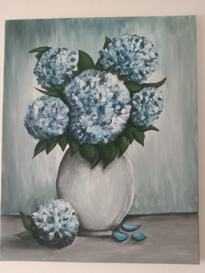 This is a painting of a white vase filled with light blue hydrangeas, set against a muted teal background. Several hydrangeas are clustered within the vase, with green leaves visible among them. To the left of the vase, a single hydrangea bloom rests on a gray surface, accompanied by a couple of light blue petals. The painting is displayed on a plain white wall, suggesting it is likely in a home or gallery setting, but its specific location in the city of Kielce, Poland cannot be determined from the image alone. The scene is static and depicts a still life arrangement. There are no people or animals present in the painting.