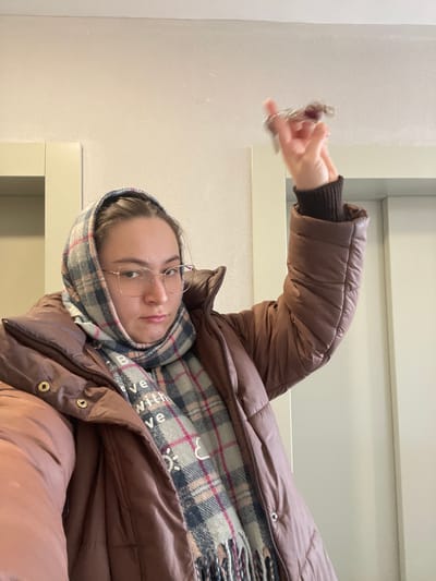 An individual is depicted indoors in Kupavna, Russia, standing in what appears to be a hallway or common area of an apartment building, indicated by two light-colored vertical door frames or elevator panels on either side of the background. The walls and ceiling are plain and light-colored. The subject is a female wearing eyeglasses, a brown puffer jacket, and a plaid scarf draped over her head and neck. The scarf features a pattern of light blue/grey, dark blue, pink, and white, with text "BELIEVE WITH LOVE" and small heart and sun icons. Her right arm is raised, bent at the elbow, and her hand holds a set of keys with multiple keyrings, one appearing dark and textured, the other metallic. The keys are slightly blurred due to motion. The subject maintains a neutral to slightly stern facial expression.