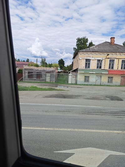 The image depicts a street scene in Едрово, Russia, on a cloudy day. The perspective is from inside a vehicle, looking out through the window, which frames the right side of the scene.

The main subjects are two buildings. On the left, there is a small, dilapidated wooden structure with a weathered appearance, featuring two boarded-up windows and a door. It appears to be abandoned or in disrepair. To the right of this, and slightly further back, stands a larger, two-story building. The upper floor is painted a pale yellow, with multiple windows and decorative architectural details. The lower floor is clad in horizontal planks painted in sections of green, yellow, and blue, with a red awning-like element on the right. This building also appears aged, with peeling paint and some visible damage. A brick chimney rises from the roof of this building.

In front of the buildings is a paved area, likely a road or a parking lot, which appears wet in places, suggesting recent rain. The road has tire marks and a faded white line. A large white arrow, pointing to the right, is painted on the road in the foreground, indicating a directional lane. Beyond the buildings, trees and power lines are visible, with a cloudy sky overhead. The overall impression is of a rural or small-town setting, possibly in a state of gradual decline or undergoing transition, given the contrasting conditions of the two buildings. The weather is overcast, but there are patches of blue sky and distinct white clouds, suggesting a dynamic weather pattern. No people are visible in the frame. There is no visible text.