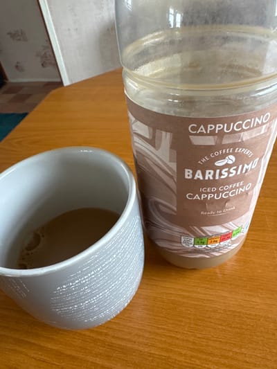 The scene is situated indoors, specifically in Fowlis Easter, United Kingdom. No individuals are present. The primary subjects are a light grey ceramic mug and a clear plastic bottle, both resting on a smooth, light brown wooden surface. The mug, positioned in the lower-left, contains a light brown liquid with a thin layer of foam, consistent with coffee. Its exterior features a subtle, textured white pattern. To the right, a mostly empty clear plastic bottle stands upright. The bottle's brown label is partially visible, indicating "CAPPUCCINO," "THE COFFEE EXPERTS," and "BARISSIMO ICED COFFEE CAPPUCCINO Ready to Drink," along with nutritional information. The liquid level inside the bottle is low, suggesting a significant portion has been consumed or poured out. In the blurred background, a wall with light-colored, patterned wallpaper and a glimpse of a dark teal floor element are visible.