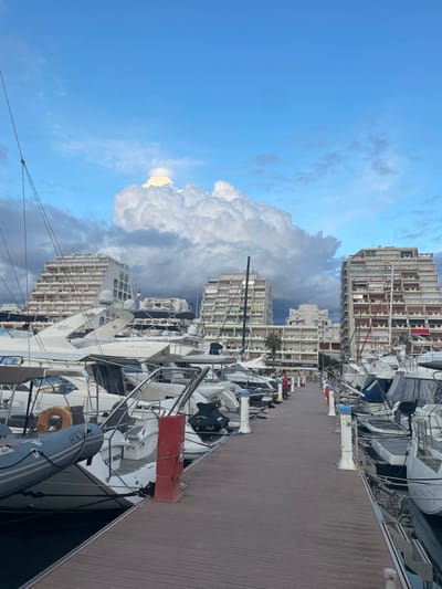 The image depicts a marina in La Grande-Motte, France, under a partly cloudy sky. A large, bright white cumulus cloud dominates the upper central portion of the sky, with areas of clear blue visible, particularly to the right, and darker grey clouds along the horizons.
A wooden boardwalk-style dock extends into the middle distance, bordered on both sides by moored watercraft. The boats primarily consist of white-hulled motor yachts and sailing boats of varying sizes, exhibiting details such as masts, rigging, railings, and covers. Visible identifiers include "AZIMUT" on a large yacht in the mid-left and "LE NOA" on a smaller boat in the lower-left. A grey inflatable dinghy with an orange lifebuoy is secured in the lower-left foreground. White and blue capped mooring posts are regularly spaced along the dock edges, and red fire safety equipment boxes are present, notably in the mid-left foreground.
In the background, distinctive multi-story residential buildings with stepped, pyramid-like architectures, characteristic of La Grande-Motte, are visible. Two prominent light-colored buildings with numerous balconies flank the central perspective, with additional structures visible further down the marina. A single palm tree is observed between the central background buildings. No individuals are visible in the scene. The overall lighting suggests daylight conditions without harsh shadows.