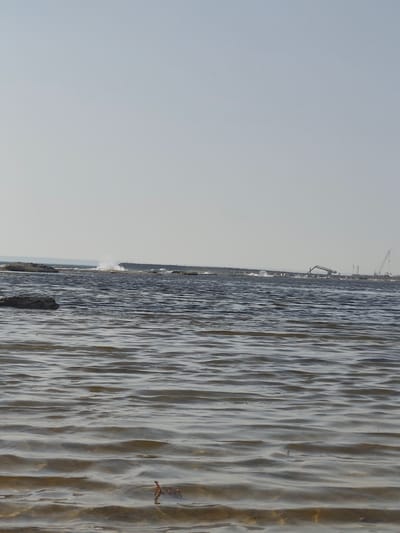 The image depicts a body of water with gentle ripples, likely a lake or sea. In the background, a breakwater or pier is visible, with some construction activity occurring. Two cranes and what appears to be an excavator are present on the breakwater, suggesting ongoing building or maintenance work. Waves are breaking against the breakwater, creating white spray. The sky is clear and pale blue, indicating fair weather. Rocks are scattered in the foreground, some submerged in the water. The scene is likely located along the coast of the Caspian Sea in the vicinity of Saiyn, Kazakhstan, an area known for its oil and gas industry and associated infrastructure development.