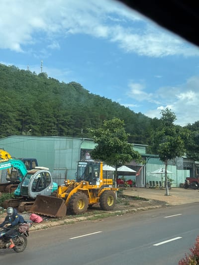 The image shows a street scene in Pleiku, Vietnam, with several construction vehicles parked along the roadside. A yellow front-end loader is prominent in the foreground, with its bucket lowered. Behind it, a light blue excavator and a teal-colored mini-excavator are visible. A motorcycle rider, wearing a helmet and carrying bags, is parked next to the excavators. The scene is set against a backdrop of a lush green hillside covered in dense trees. Buildings with green corrugated metal roofs are visible to the left, suggesting a semi-urban or industrial area. The sky is blue with scattered white clouds, indicating daylight. The road is paved with a white line marking the center. Visible text on one of the excavators reads "NAM PHÚ QUÝ" and a phone number "0905237595". The overall impression is one of a working area where heavy machinery is stored or used.